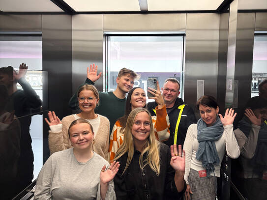 Leute stehen im Fahrstuhl und winken in die Kamera